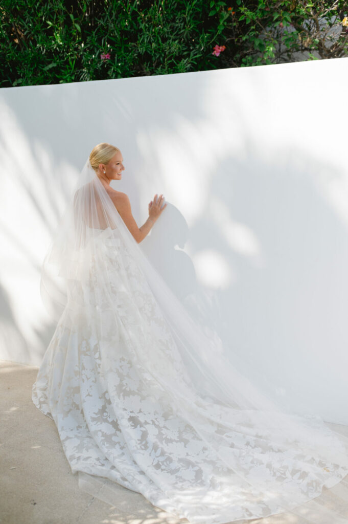 Bride in floral gown smiles by sunlit wall, greenery above.