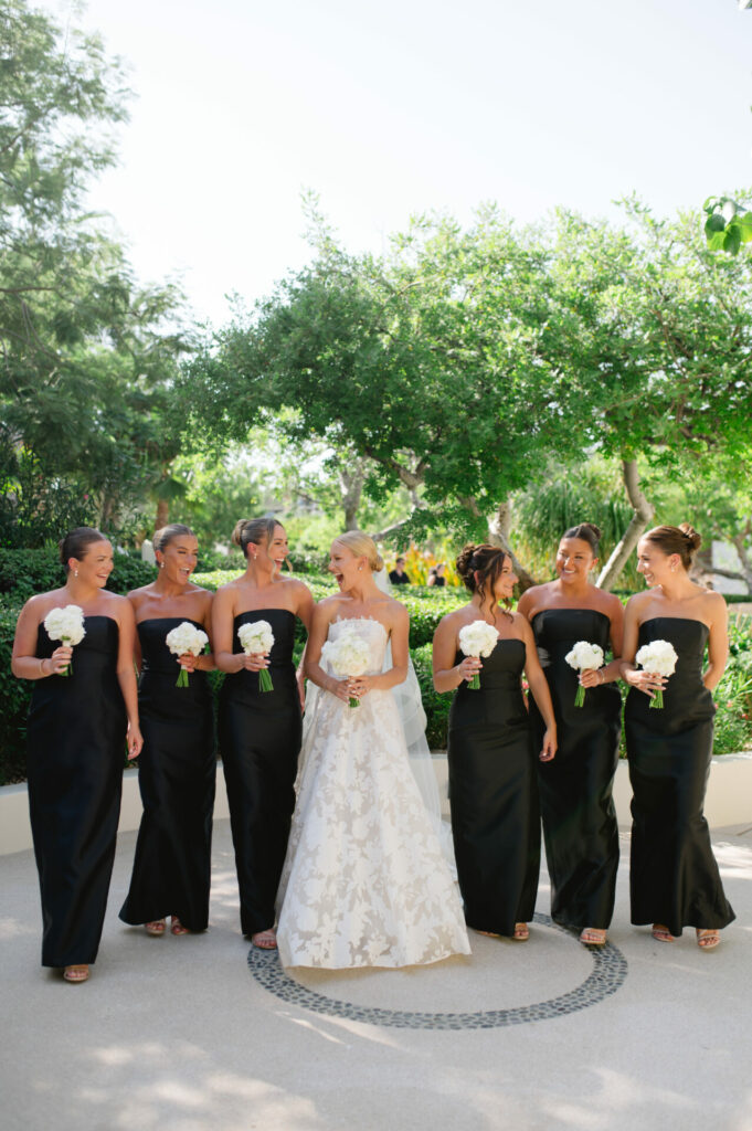 Bride in white, six bridesmaids in black, smiling outdoors.