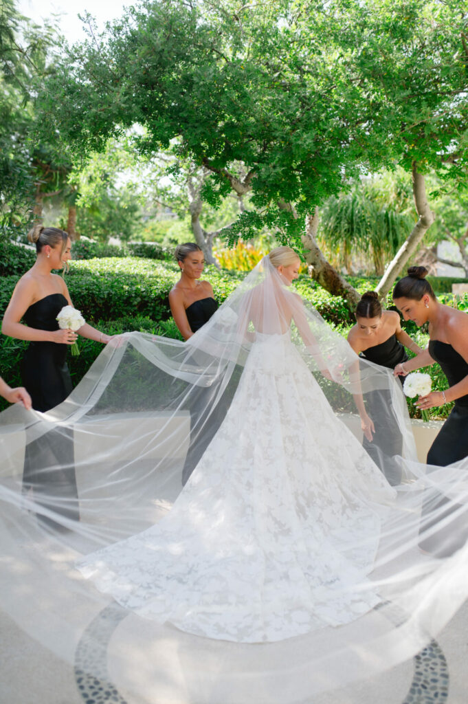 Bride in couture gown, bridesmaids fix veil outdoors.