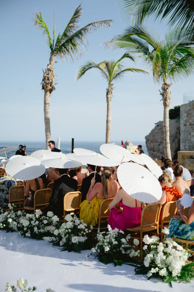 Beach wedding guests with parasols; bride in luxury gown.