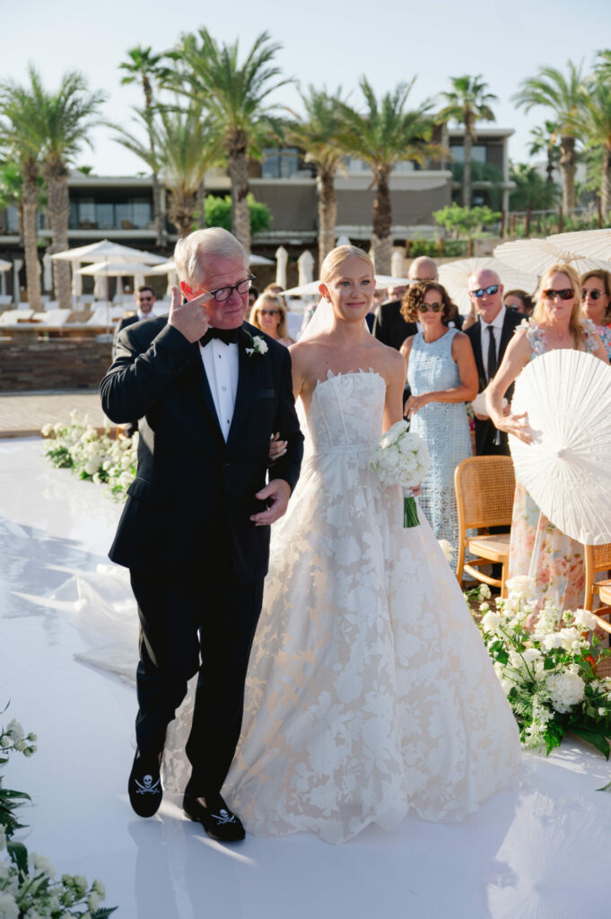 Bride in couture gown walks outdoor floral aisle, guests watch.