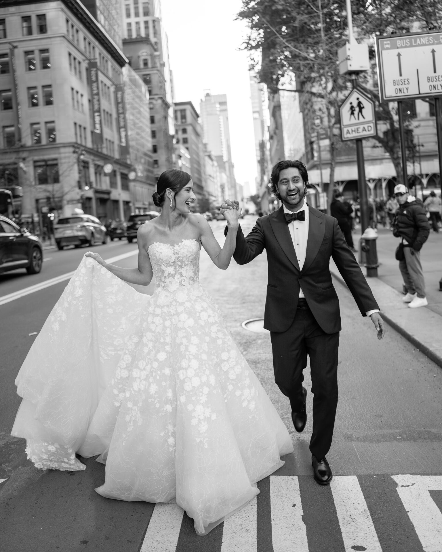 Smiling couple crosses city street in wedding attire.