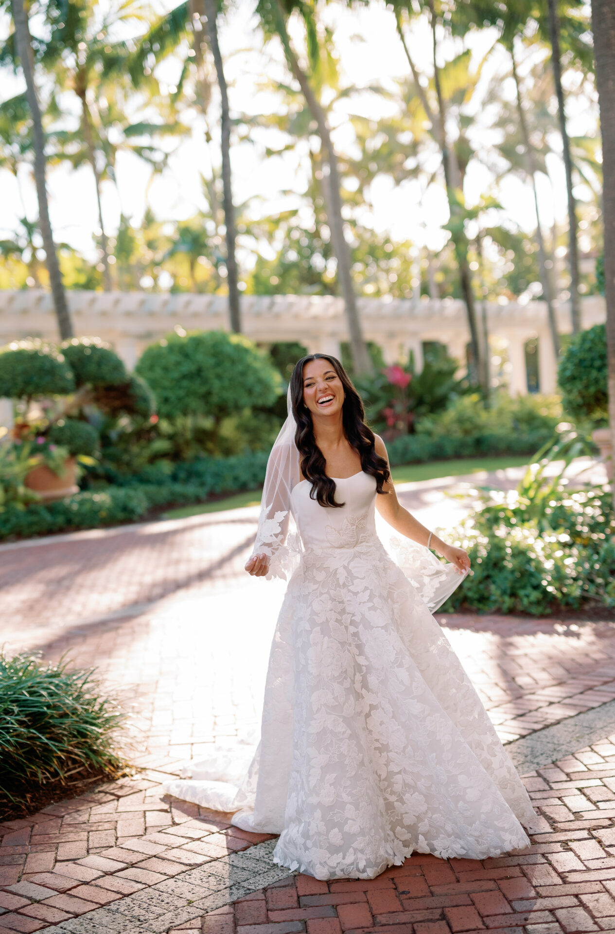 Smiling bride in white gown on sunny palm path.