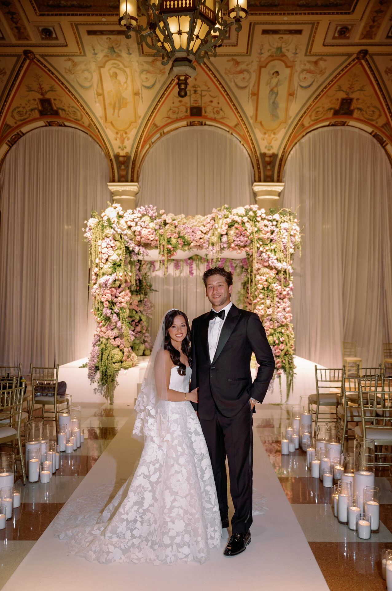 Bride and groom smiling beneath floral arch in grand hall.