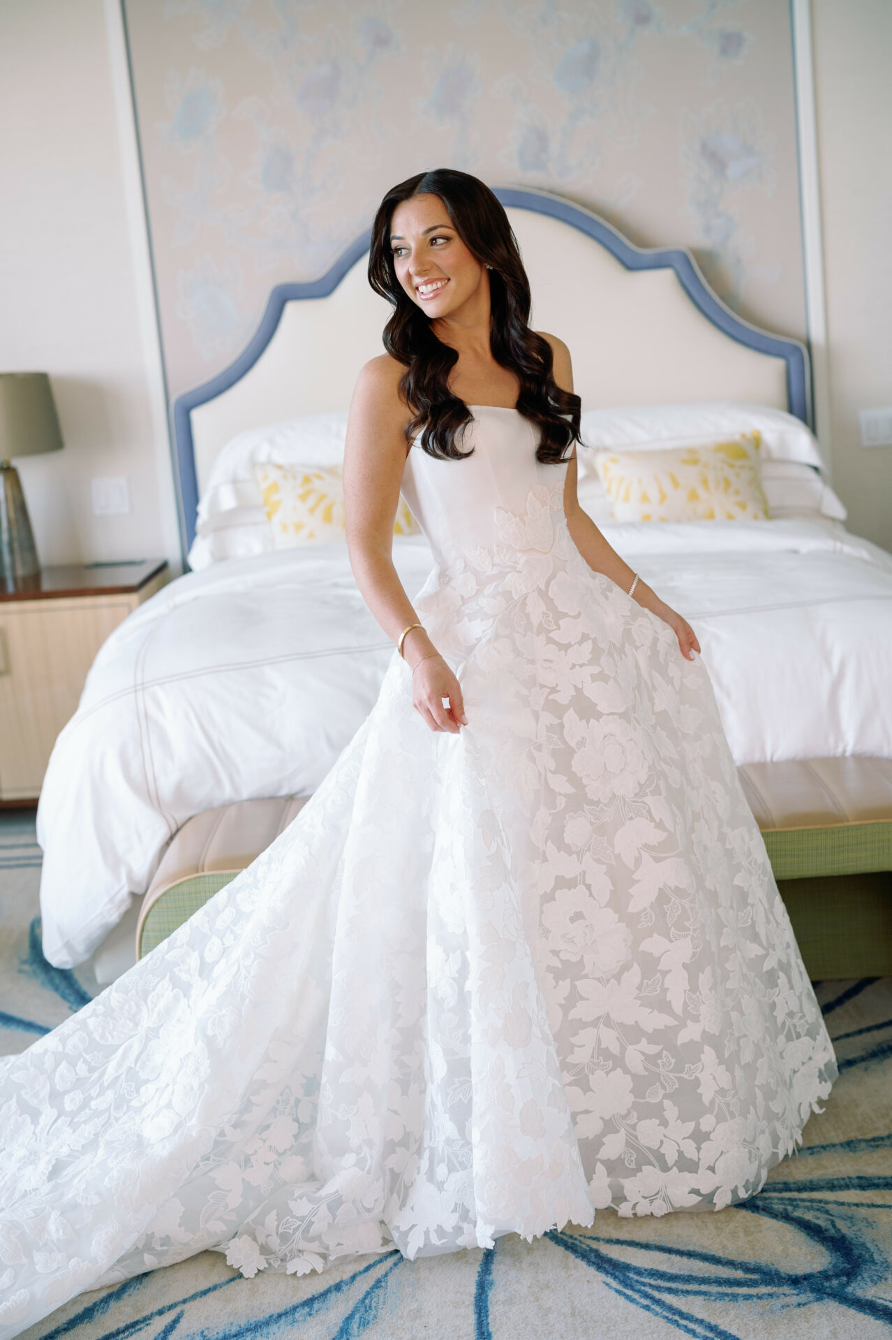 Smiling woman in strapless floral wedding dress by bed.