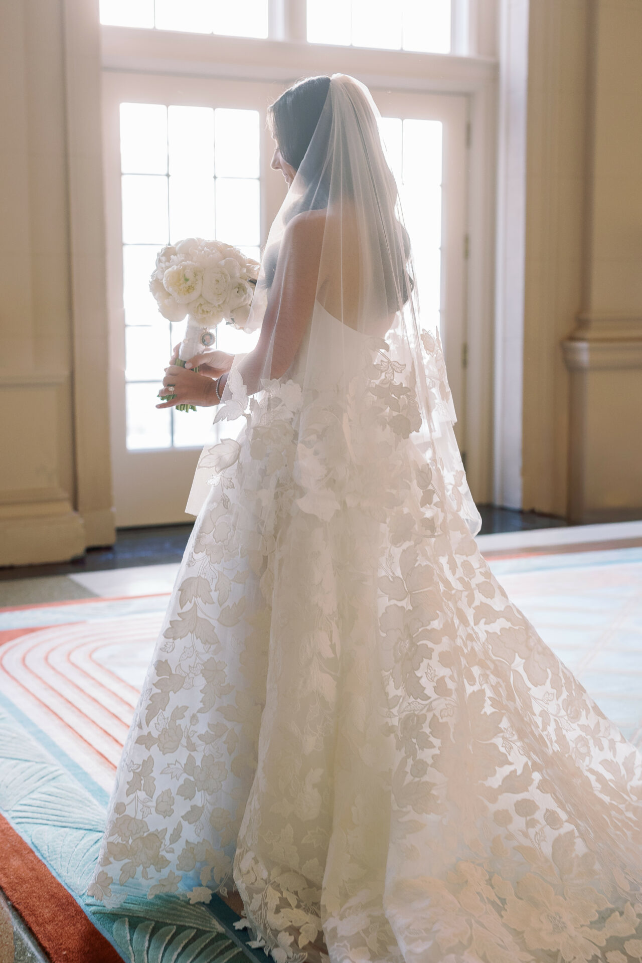 Bride in lace gown holds flowers in sunlit room.