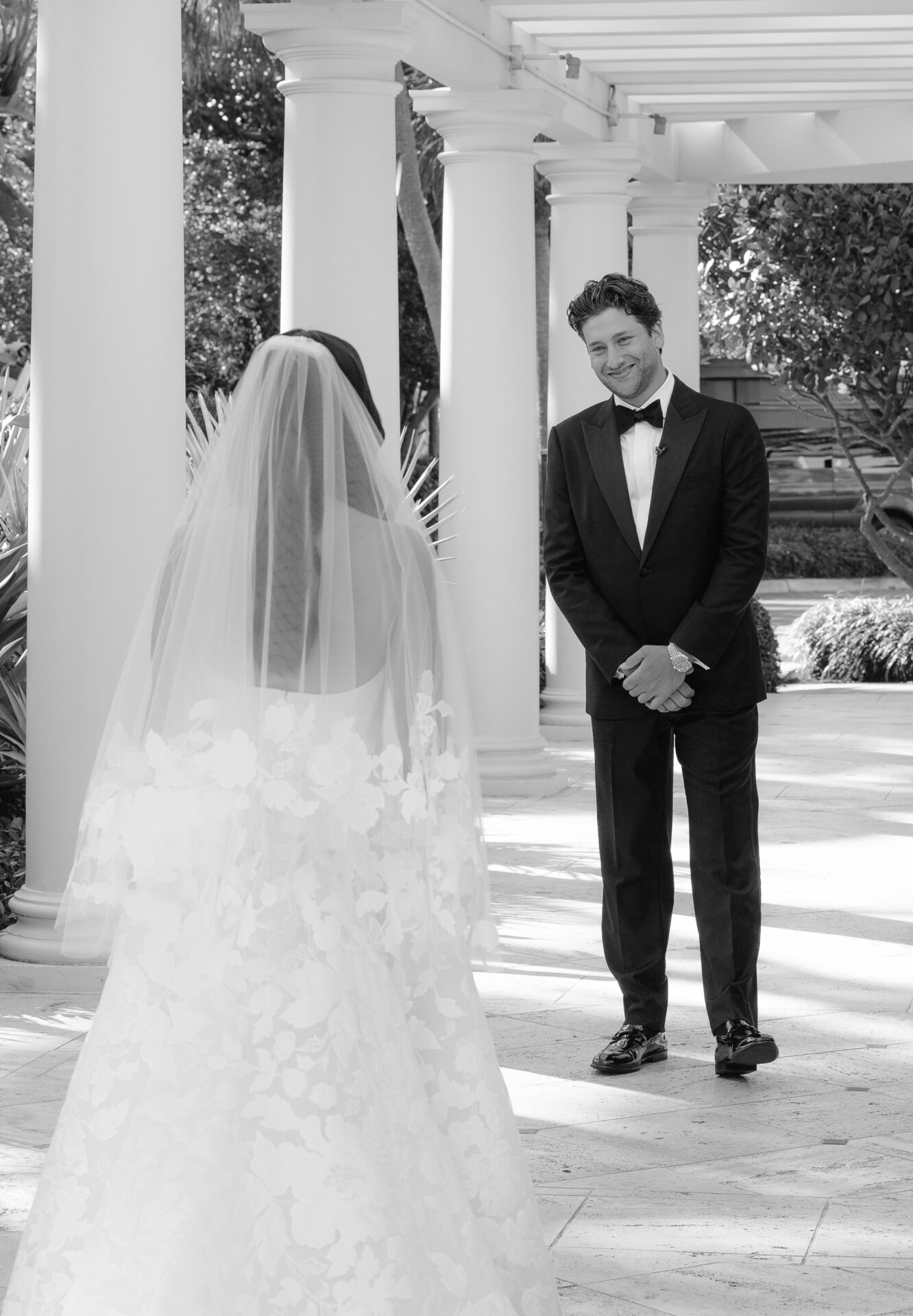 Smiling groom sees bride in floral gown outdoors.