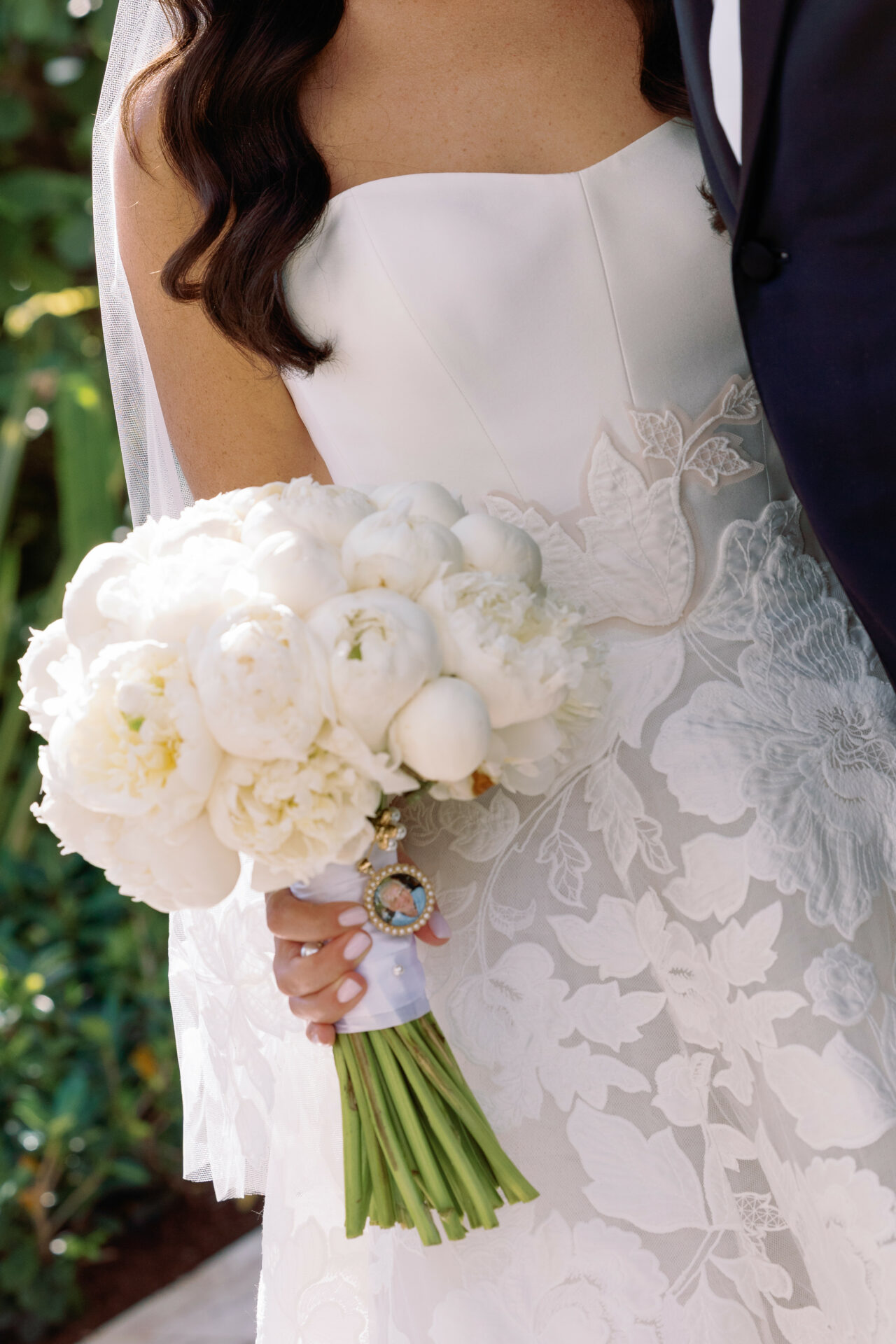 Bride in lacy gown holds peony bouquet; groom stands beside.