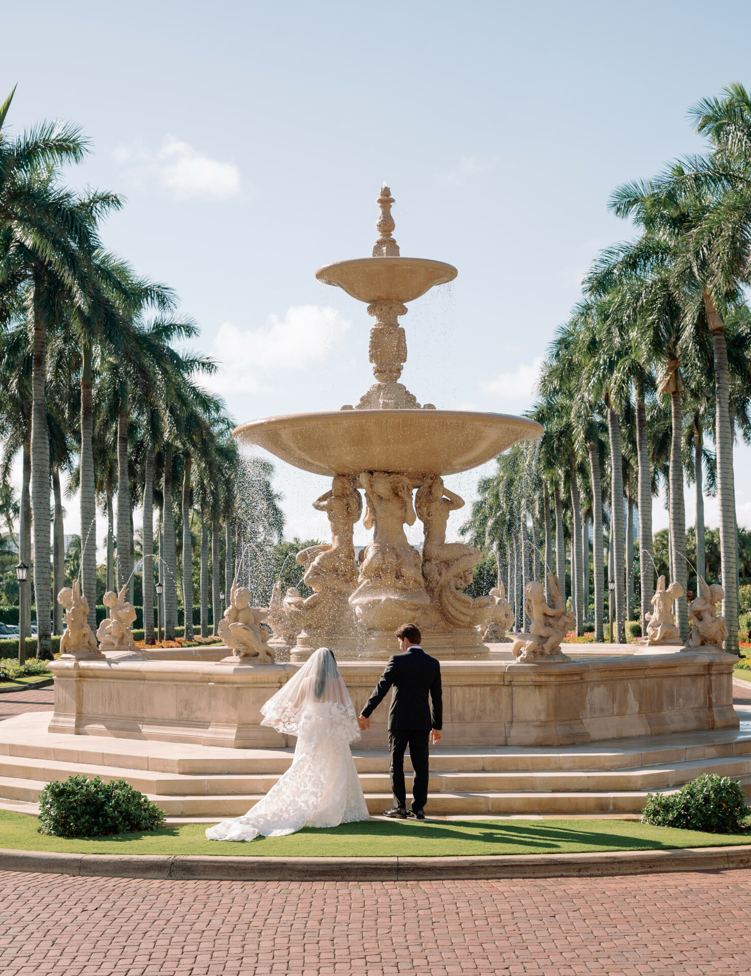 Bride and groom walk to ornate fountain under palms.