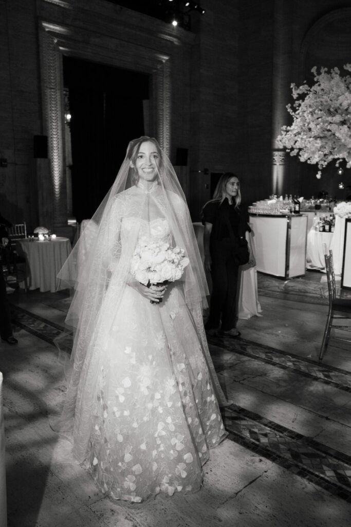 Bride in embroidered gown, holding bouquet indoors.