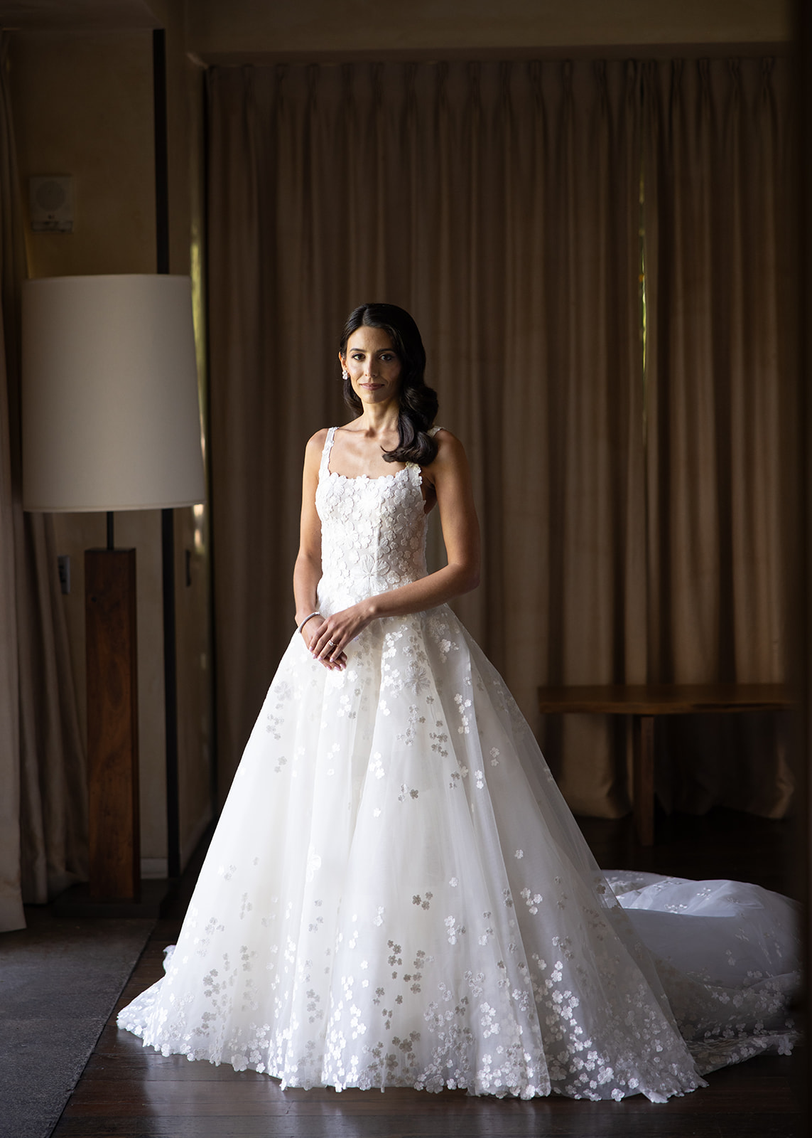 Bride in floral wedding dress stands by window.