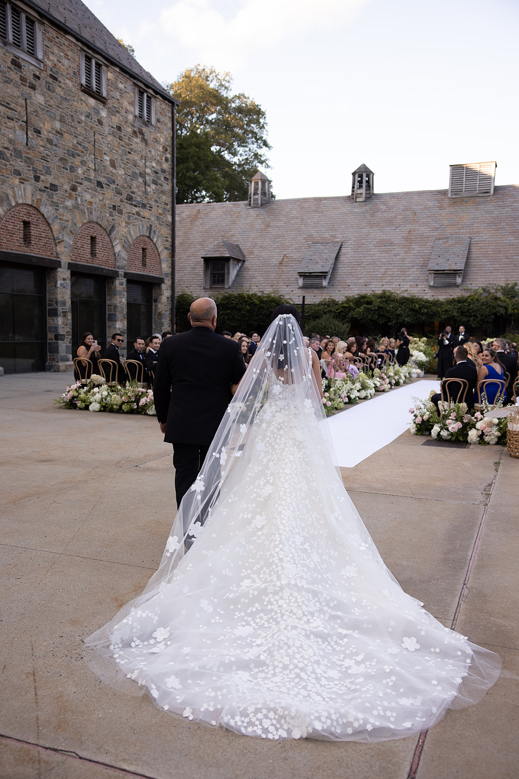 Bride in floral veil walks aisle toward guests.