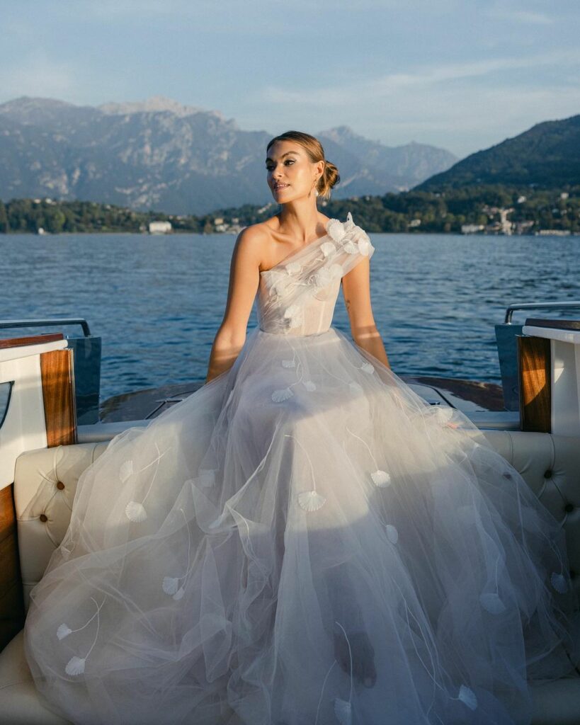 Bride in flowing white gown on boat, scenic lake.