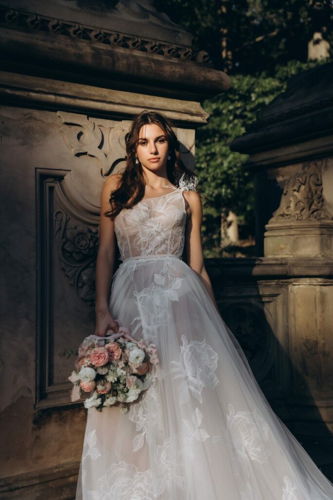 Bride in lace dress with bouquet by stone wall.