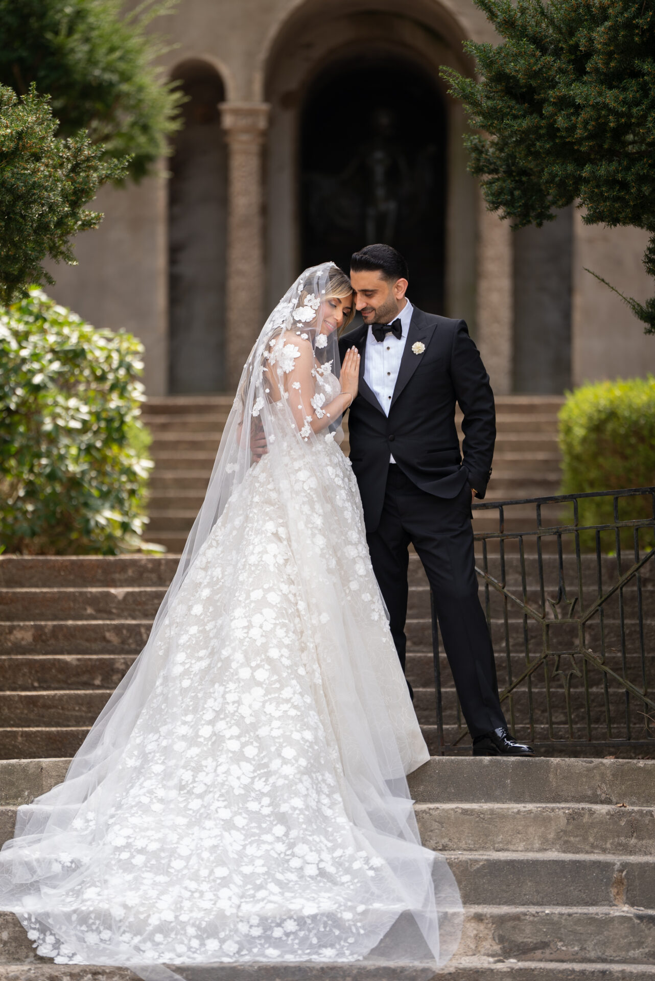 Bride in lace gown leans on groom outdoors.