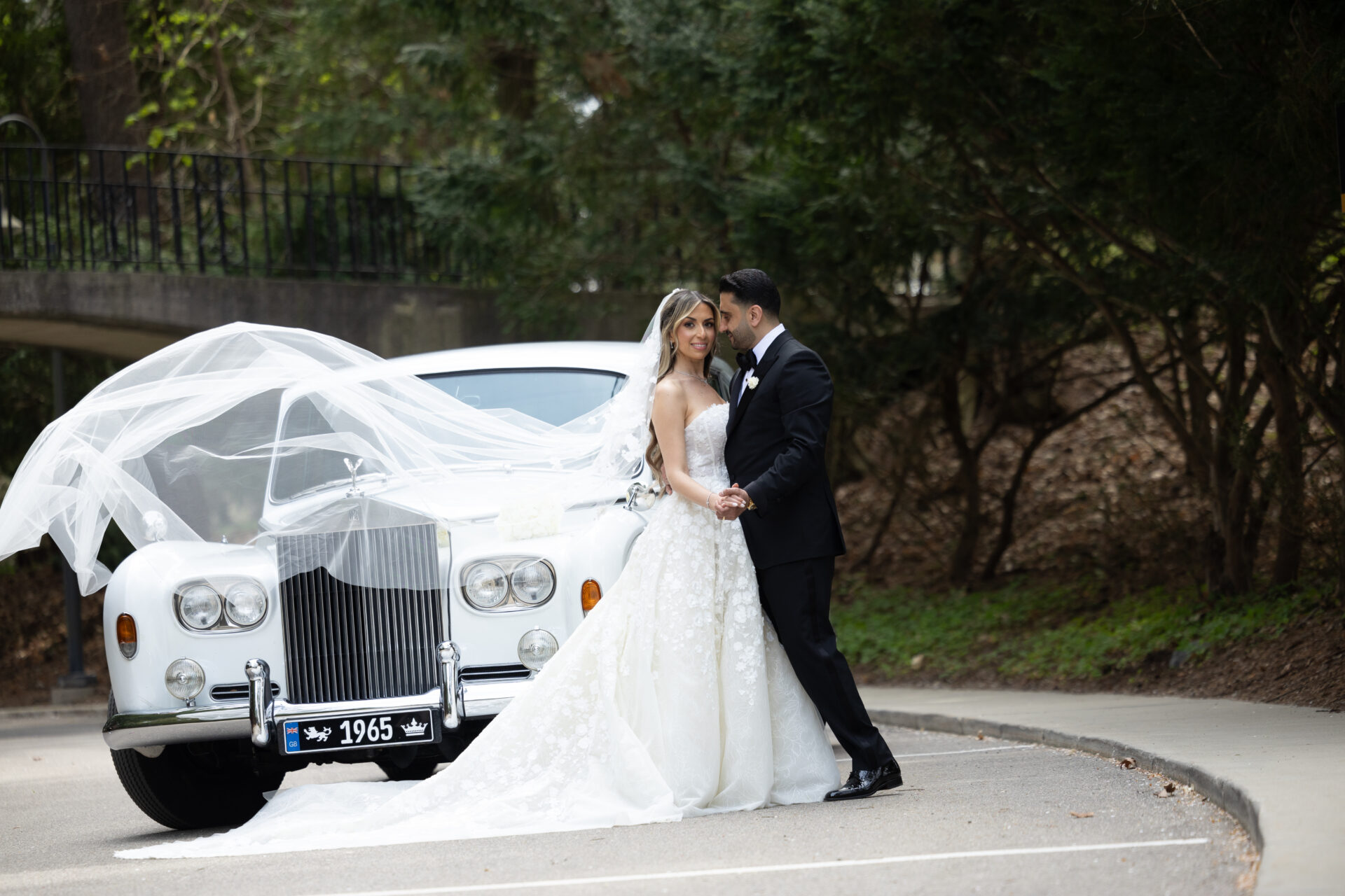 Bride and groom by vintage car, veil flowing.