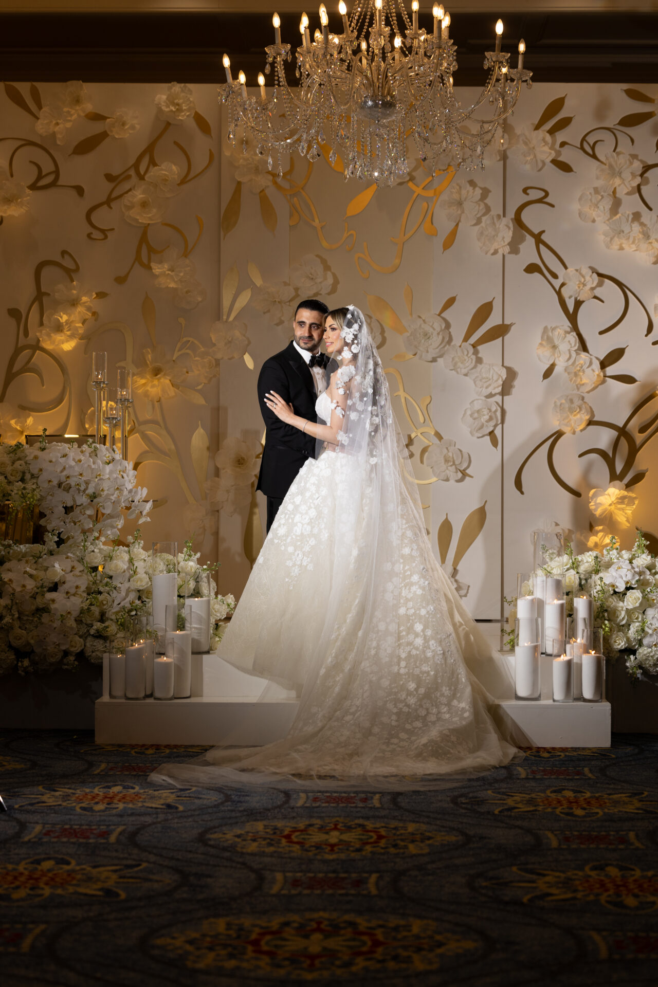 Bride and groom embrace on ornate, flower-filled stage.