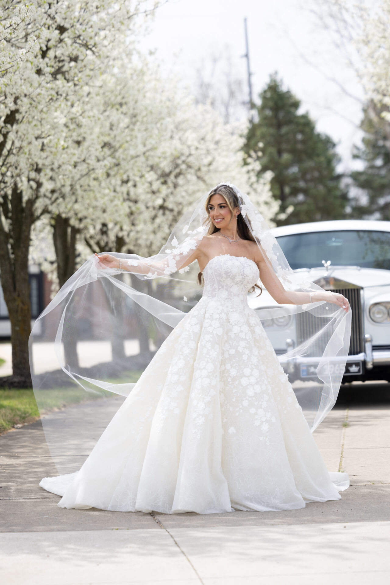 Bride in white gown poses by classic car outdoors.