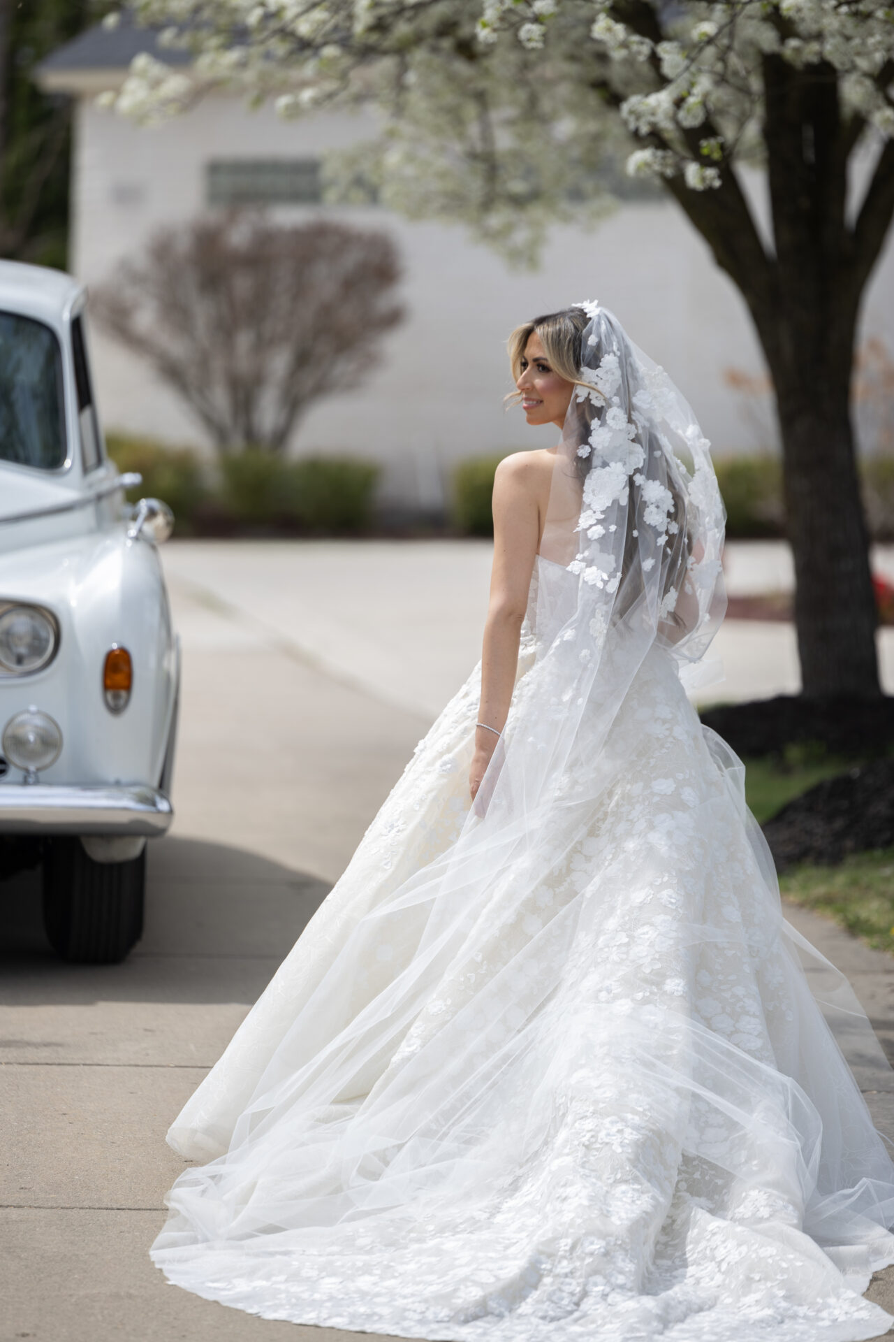 Bride in white gown by vintage car and trees.