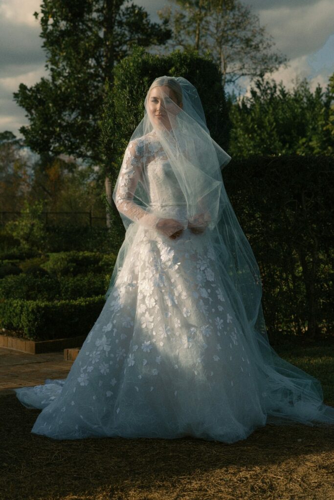 Bride in floral Mira Zwillinger gown poses in sunlit garden.
