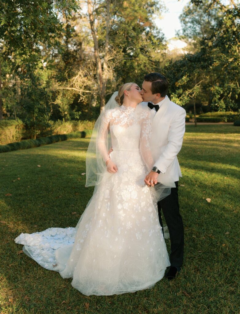 Bride in luxury gown kisses groom on tree-lined lawn.