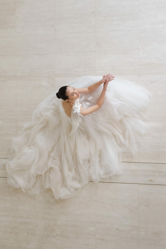 Bride in tulle gown stands on marble, looking upward.