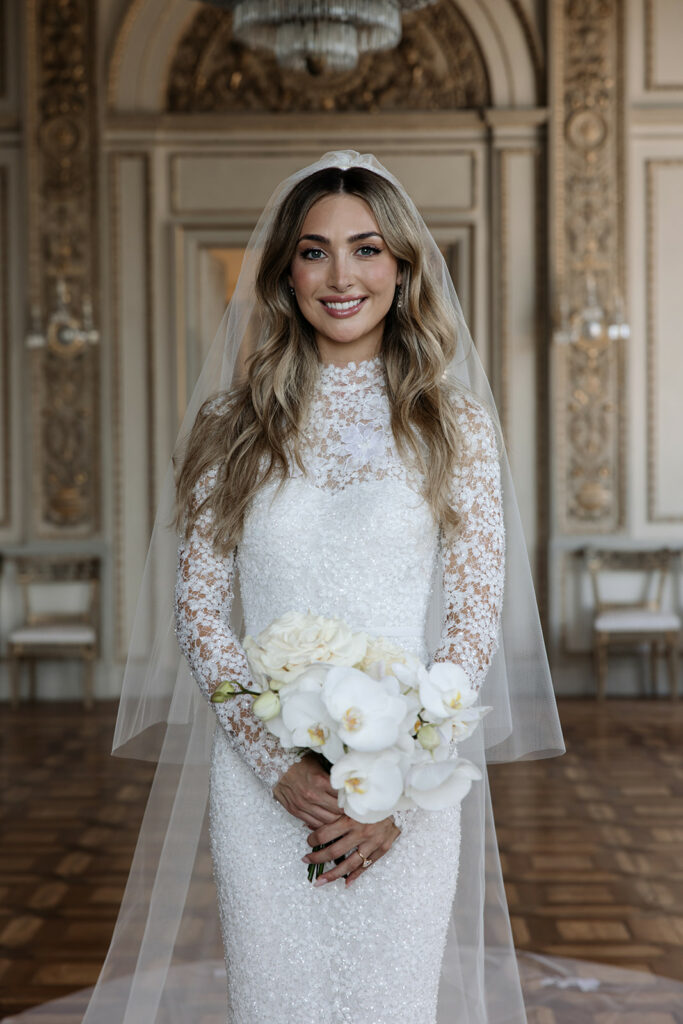 Katya Ruiz smiles in couture gown, holding white bouquet indoors.