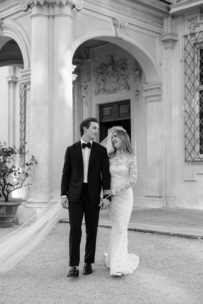 Elegant bride and groom smile outside ornate building.
