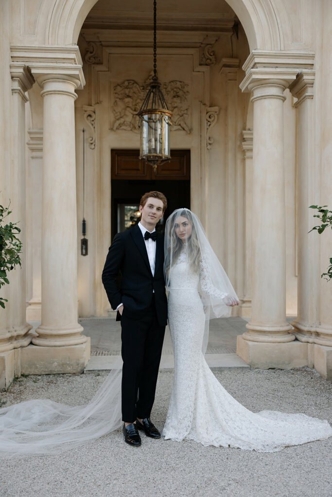 Katya Ruiz and groom pose outside grand building, elegant gown.