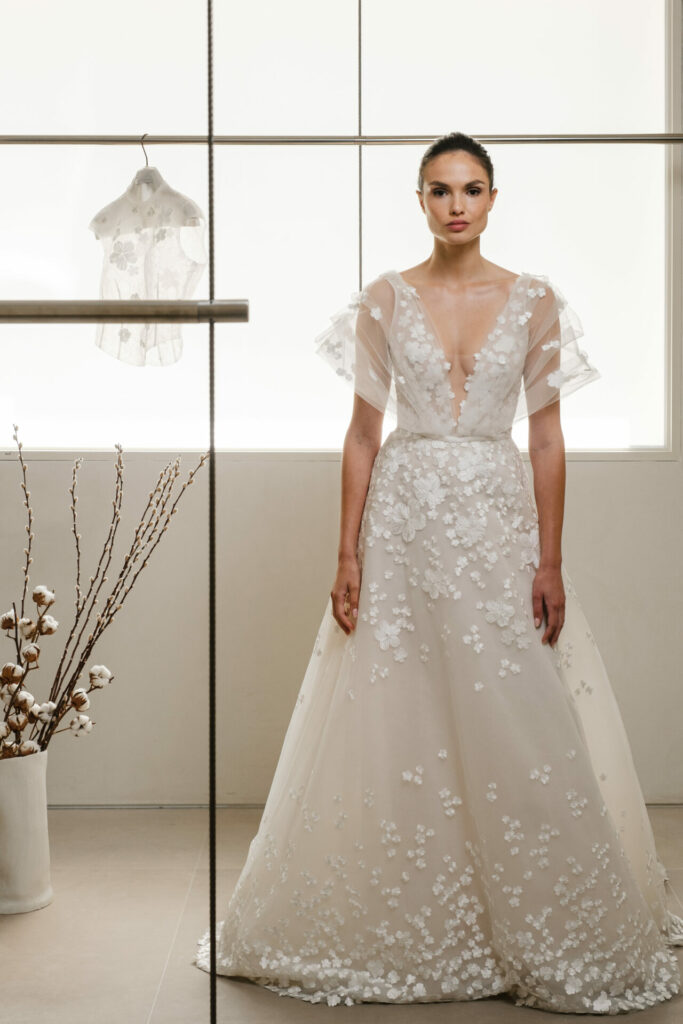 Bride in floral gown by window; bolero behind.