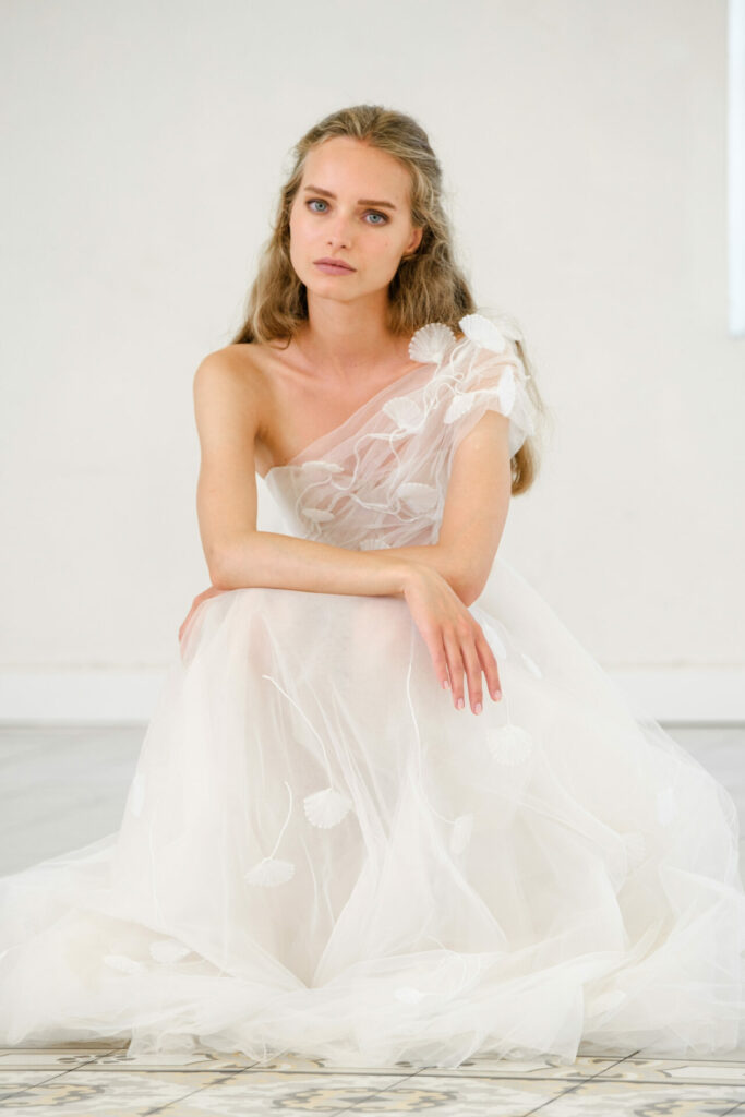Bride in flowing white gown sits calmly, posing.