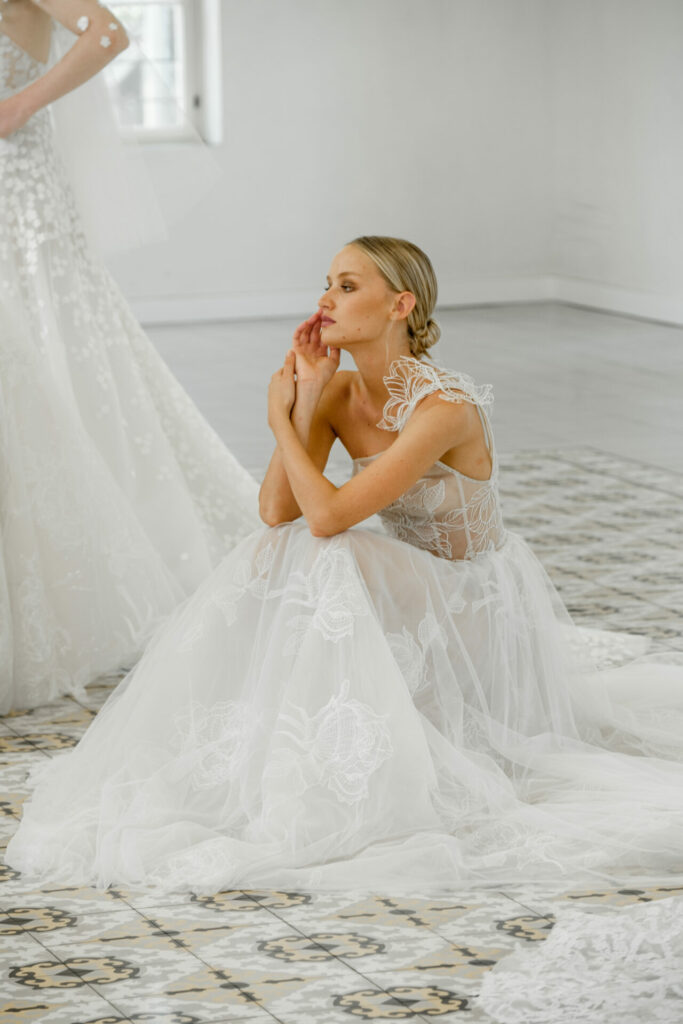 Two brides in sheer lace gowns, one seated thoughtfully.