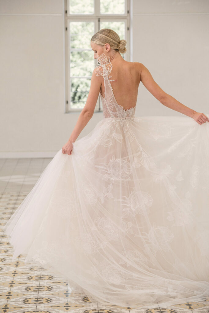 Bride in sheer lace gown poses by bright window.