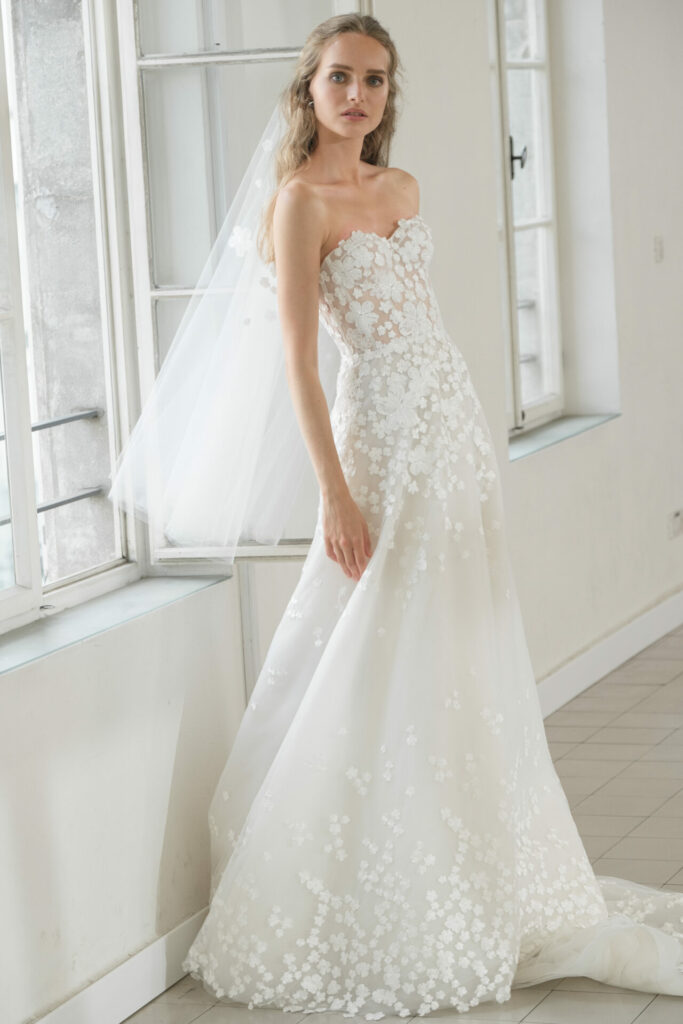 Bride in lace A-line gown stands by windows.
