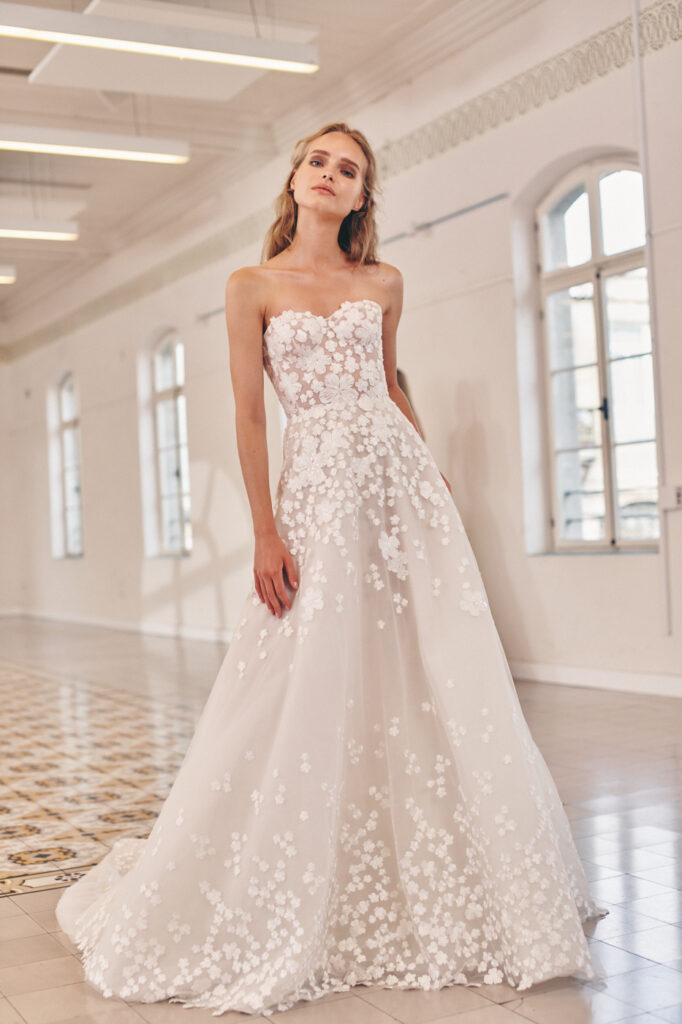 Woman in strapless lace wedding gown, large windows behind.