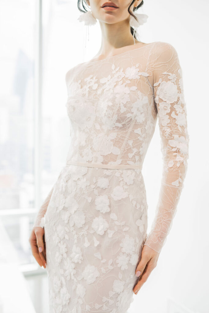 Woman in sheer floral wedding dress, sunlit room.