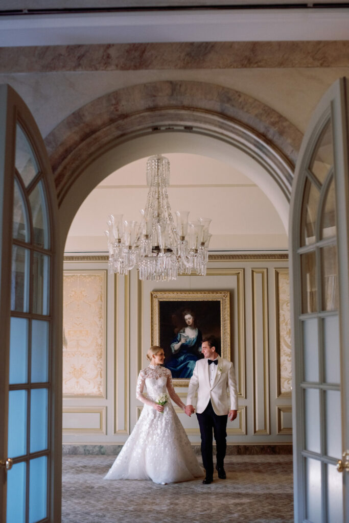 Bride and groom hold hands in ornate, luxurious room.