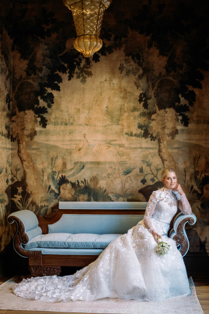 Katya Ruiz in couture gown on vintage blue couch, elegant room.