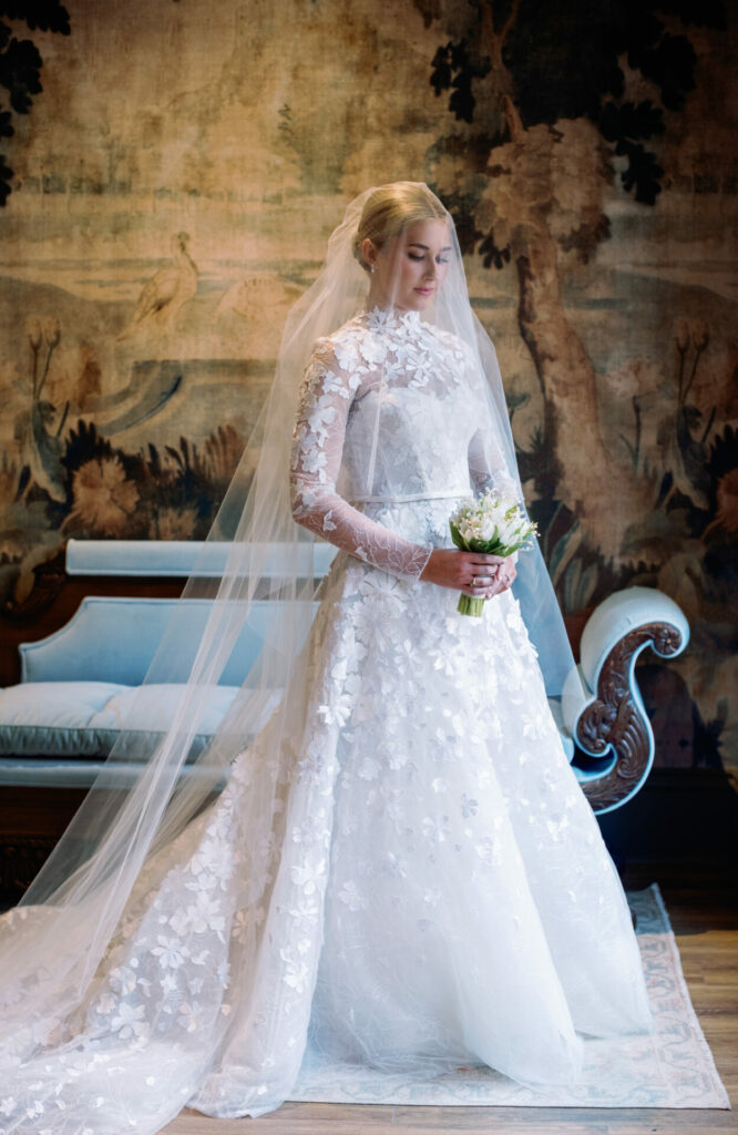 Katya Ruiz in lace gown indoors with bouquet, sofa, mural.