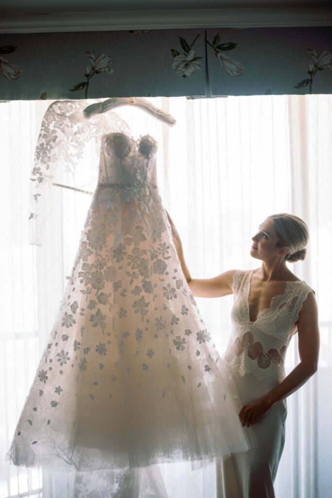 Woman in lace dress holds bridal gown by window.