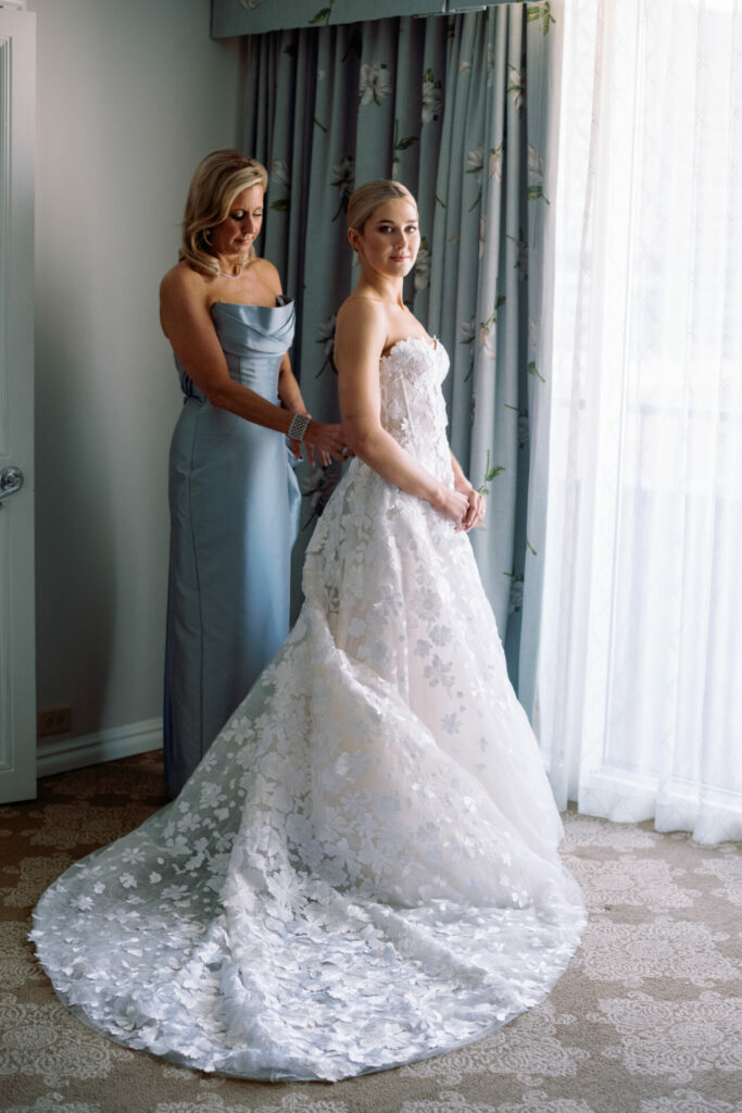 Bride in lace gown by window, dress adjusted.