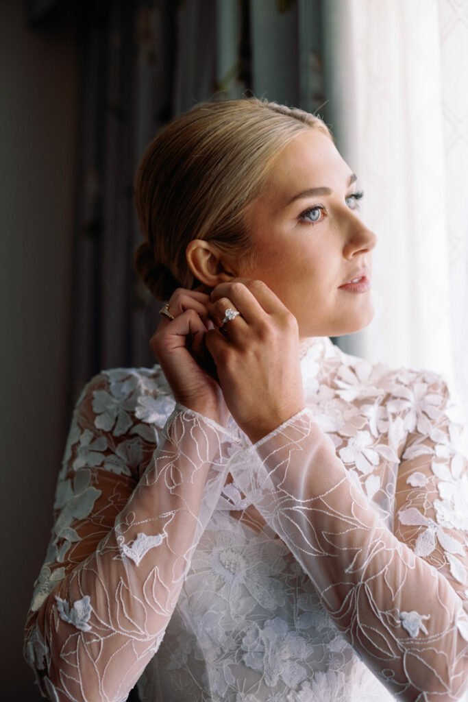 Blonde bride in lace gown adjusts earring by window.