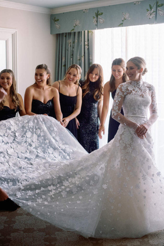 Bride in lace gown with five bridesmaids admiring train.