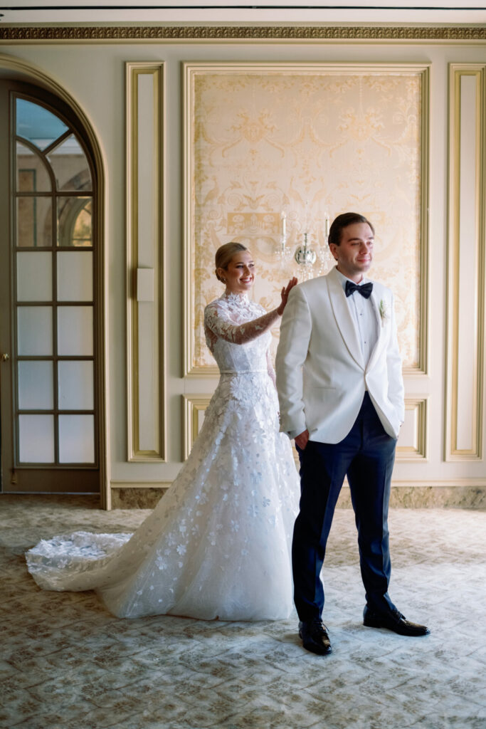 Katya Ruiz in couture gown stands behind groom for first look in an ornate, softly lit room.