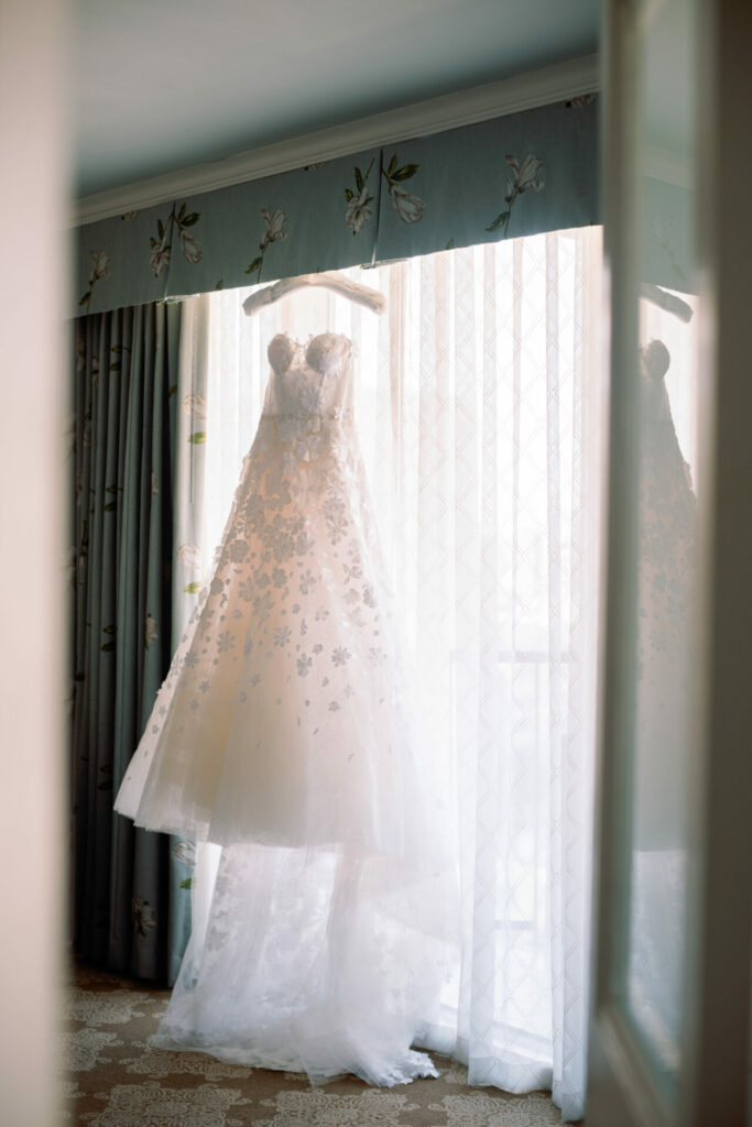 White, strapless floral gown hangs by sunlit window.