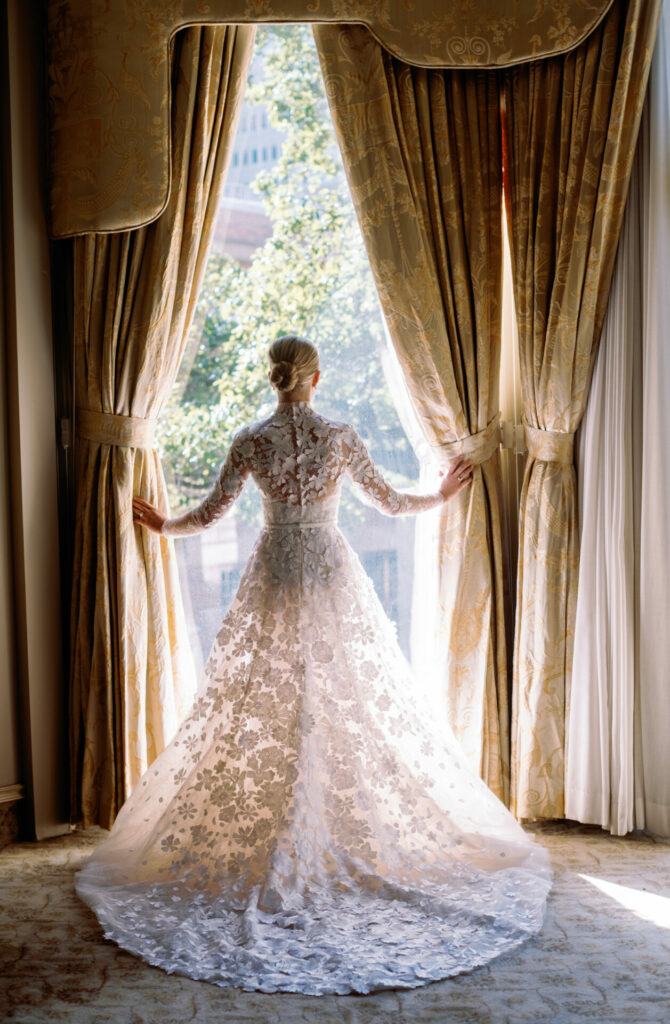 Bride in lace gown opens gold curtains to sunlight.