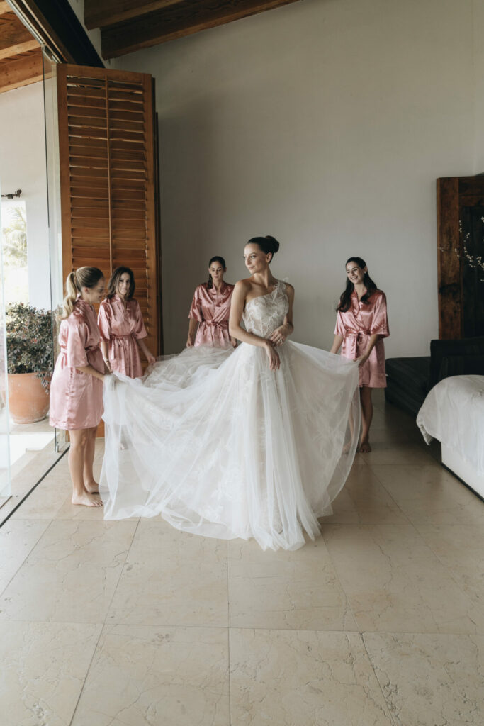 Bride in corset gown, bridesmaids adjust dress, modern room.