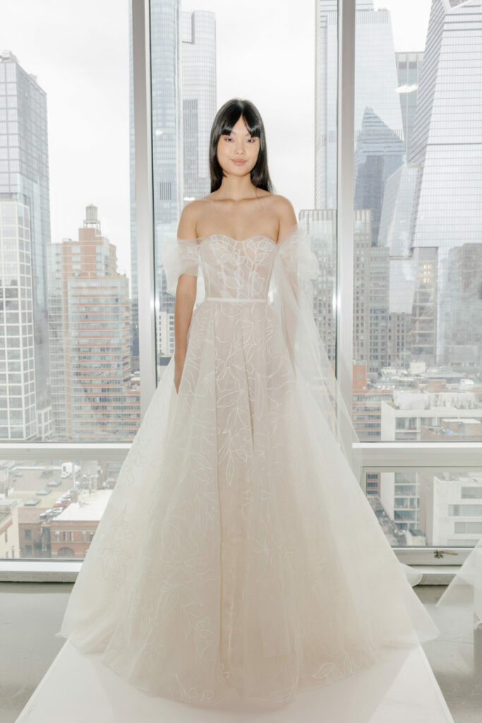 Woman in sheer white gown by windows, city view.
