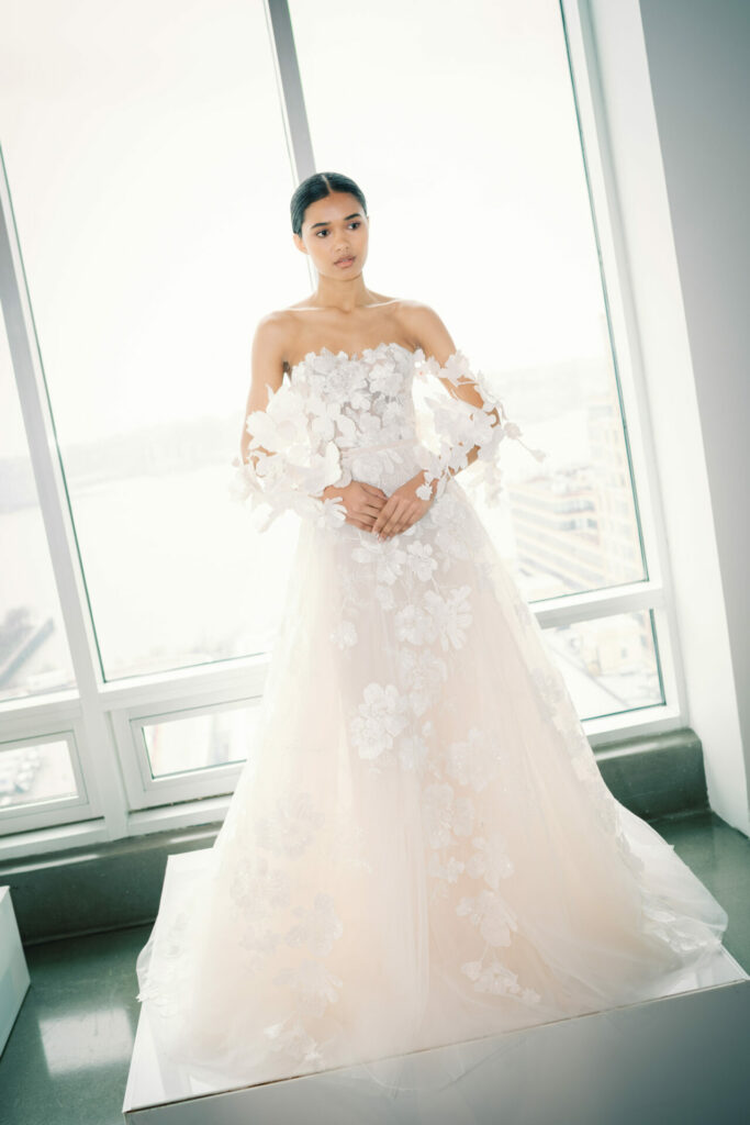 Bride in white off-shoulder gown by bright windows.