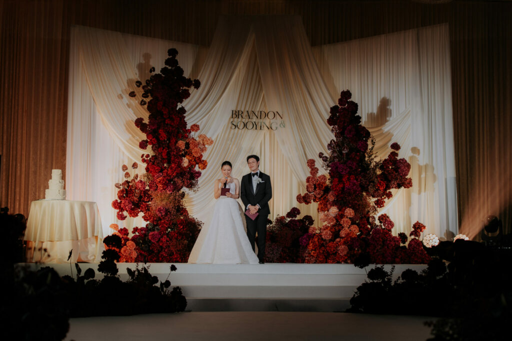 Bride and groom on floral stage, names displayed.